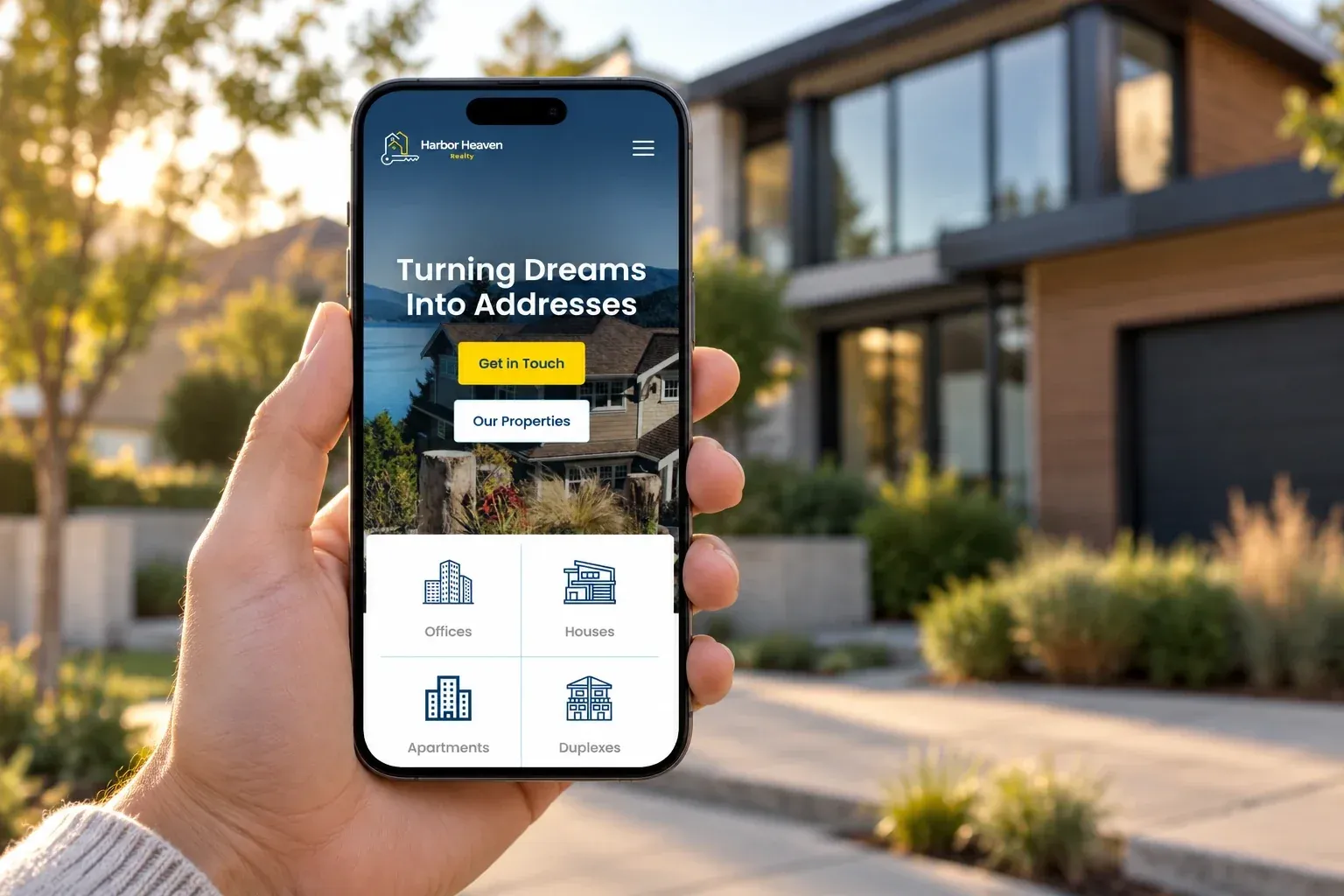 Mano che tiene uno smartphone con la homepage di Harbor Heaven Realty davanti a una casa moderna al tramonto
