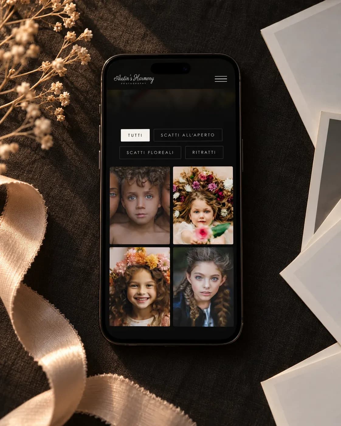Smartphone con la galleria mobile del sito, appoggiato su lino scuro tra stampe fotografiche e fiori secchi