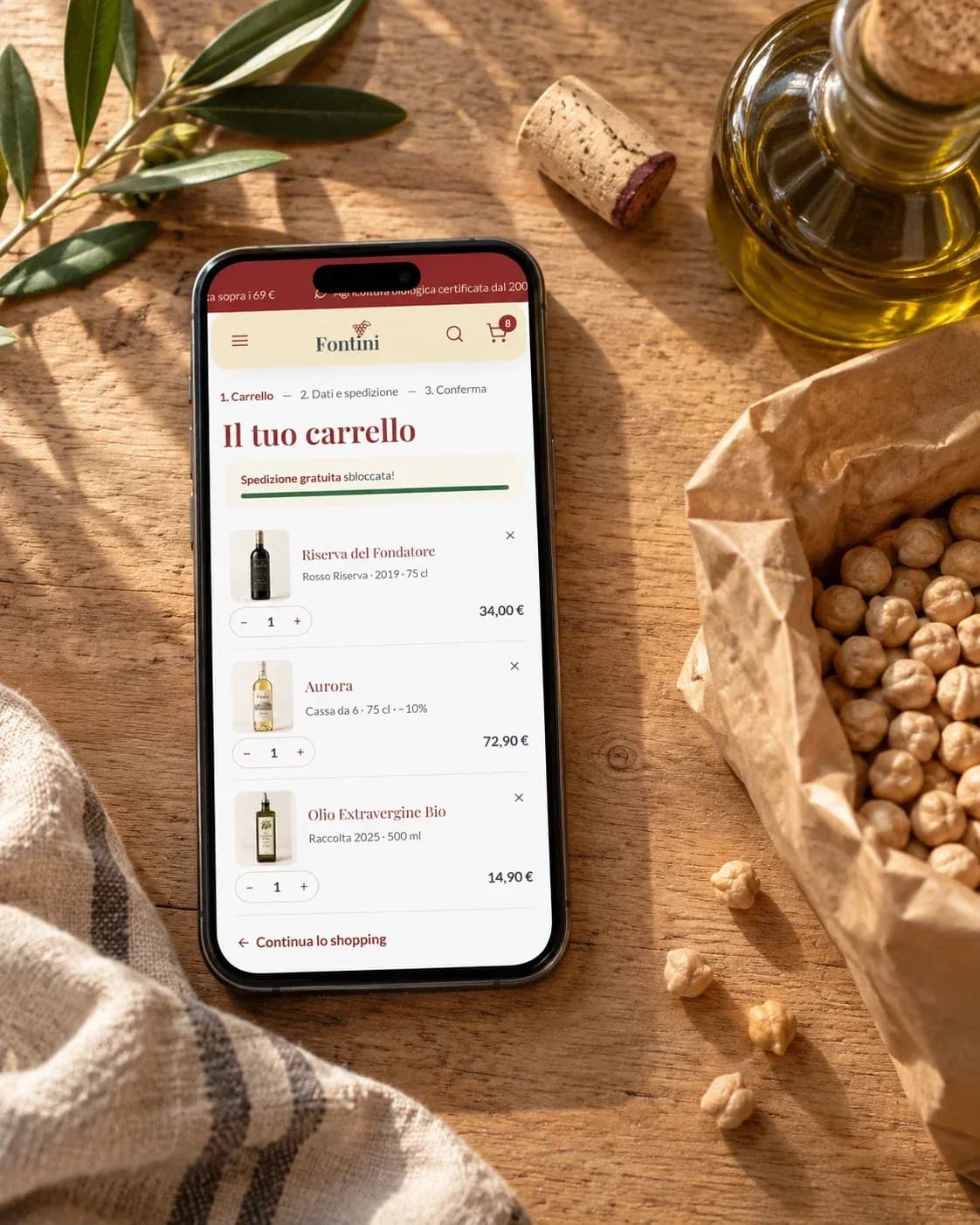 Smartphone con il carrello di Fontini su un tavolo di legno, accanto a un sacchetto di ceci, una bottiglia d'olio e un tappo di sughero