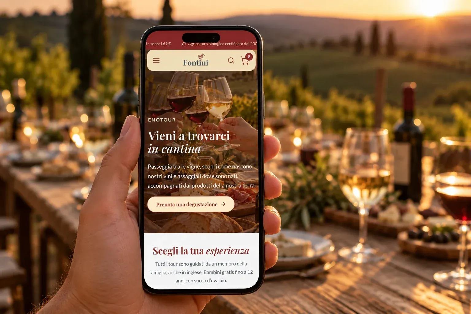 Mano che tiene uno smartphone con la pagina EnoTour di Fontini davanti a una tavola apparecchiata per una degustazione in vigna al tramonto