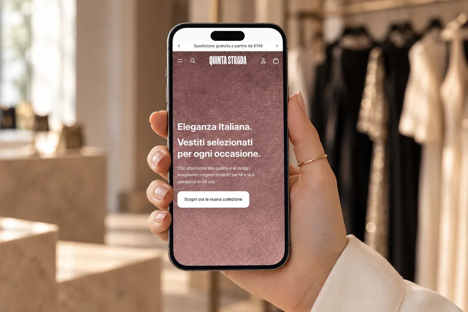 Mano che tiene uno smartphone con la homepage mobile di Quinta Strada, sullo sfondo una boutique di abiti eleganti