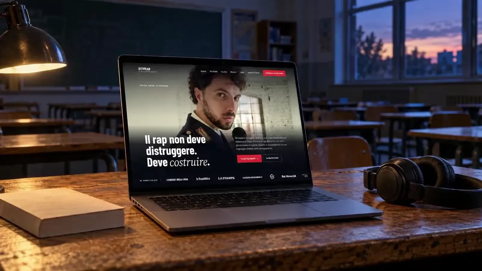 Homepage del sito di REVMAN su un portatile appoggiato sulla cattedra di un'aula scolastica al tramonto, accanto a un libro e a delle cuffie