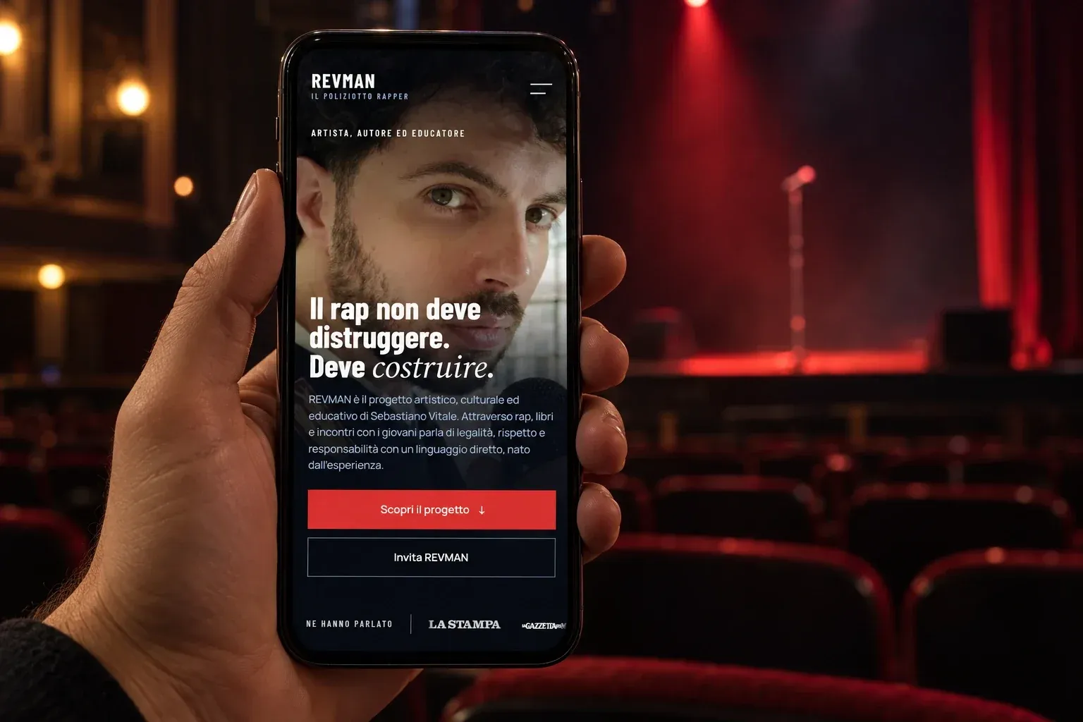 Mano che tiene uno smartphone con la homepage mobile di REVMAN, sullo sfondo un teatro con un microfono illuminato di rosso